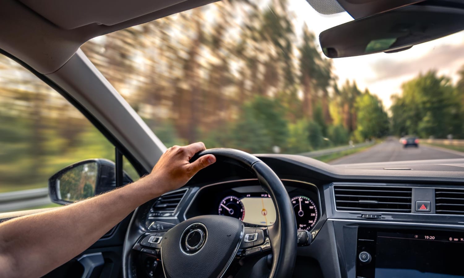 A person’s hand is on the steering wheel while driving a car down a tree-lined road. The speedometer and dashboard are visible, and the outside scenery appears blurred, indicating motion.