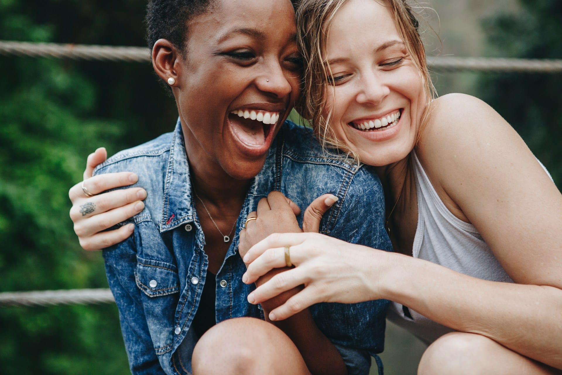 Two women sitting close together outdoors, laughing and smiling, one wearing a denim jacket and the other in a tank top, with greenery blurred in the background.